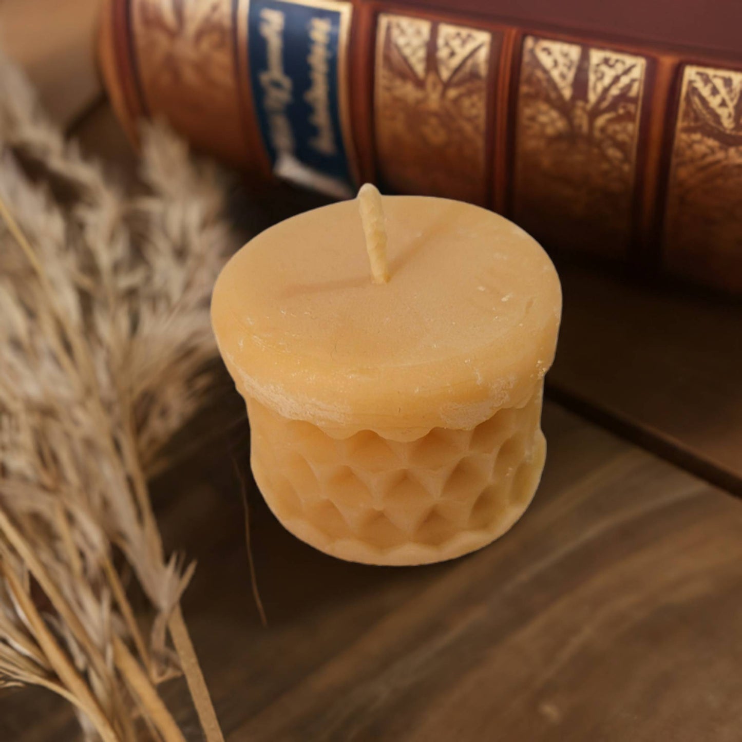 A beeswax tealight candle with a honeycomb texture, placed on a wooden surface, with dried plants and a book in the background.