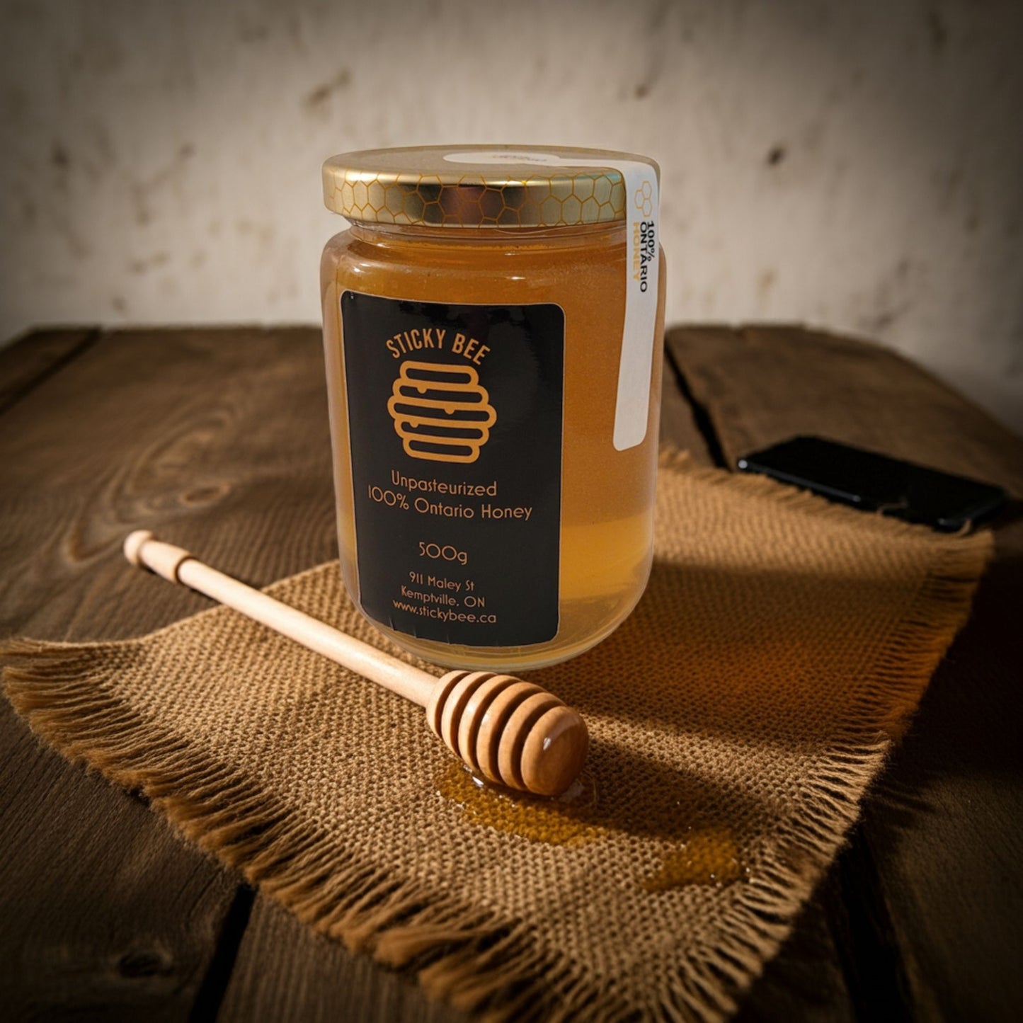Jar of honey with a honey dipper on a textured surface, featuring the 'Sticky Bee' brand.
