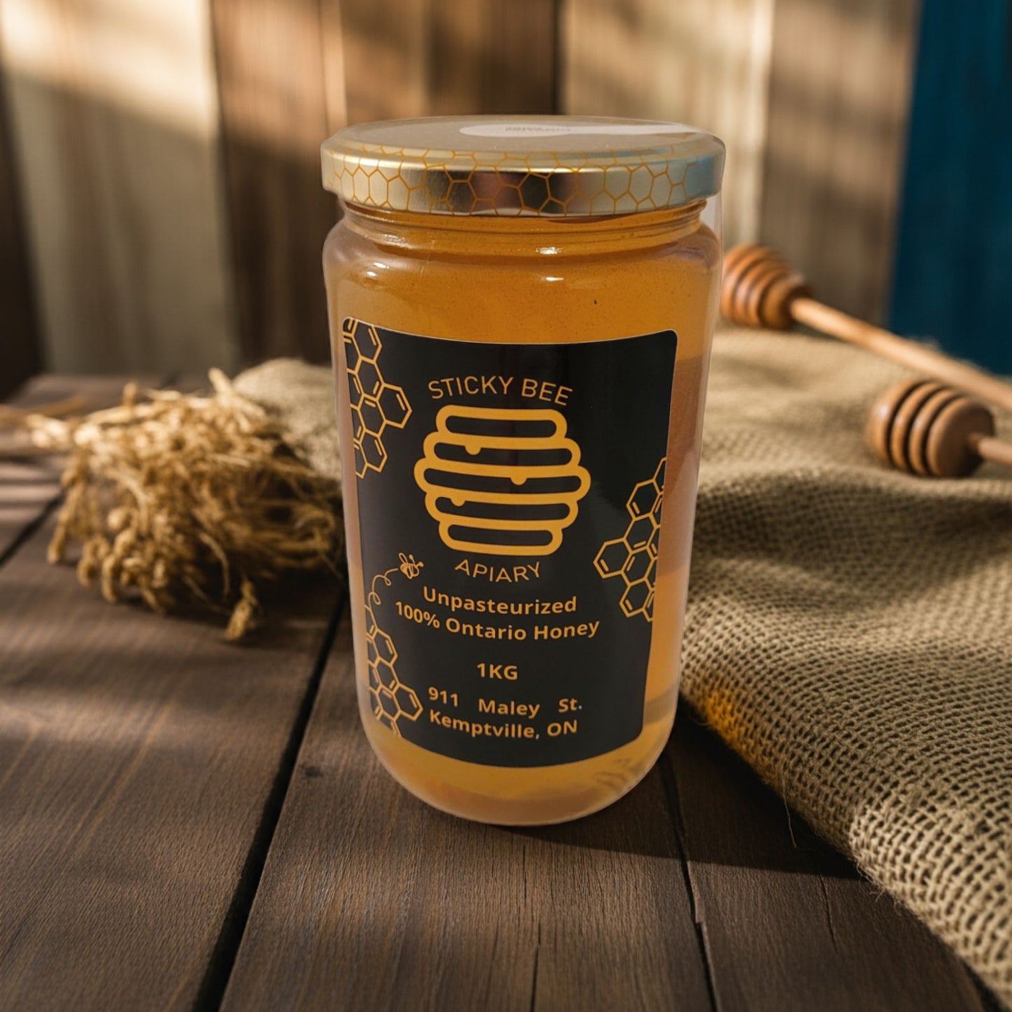 Jar of Sticky Bee Apiary honey on a wooden surface with a honeycomb and dipper in the background.