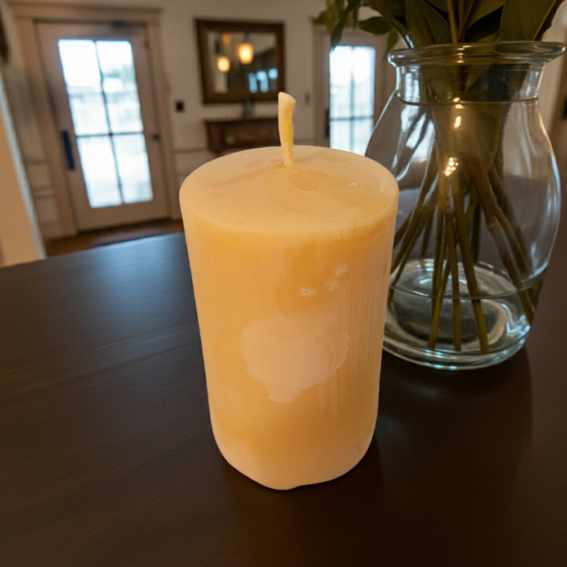 Smooth Pillar Beeswax Candle on a dark surface with a glass vase containing plants in the background.