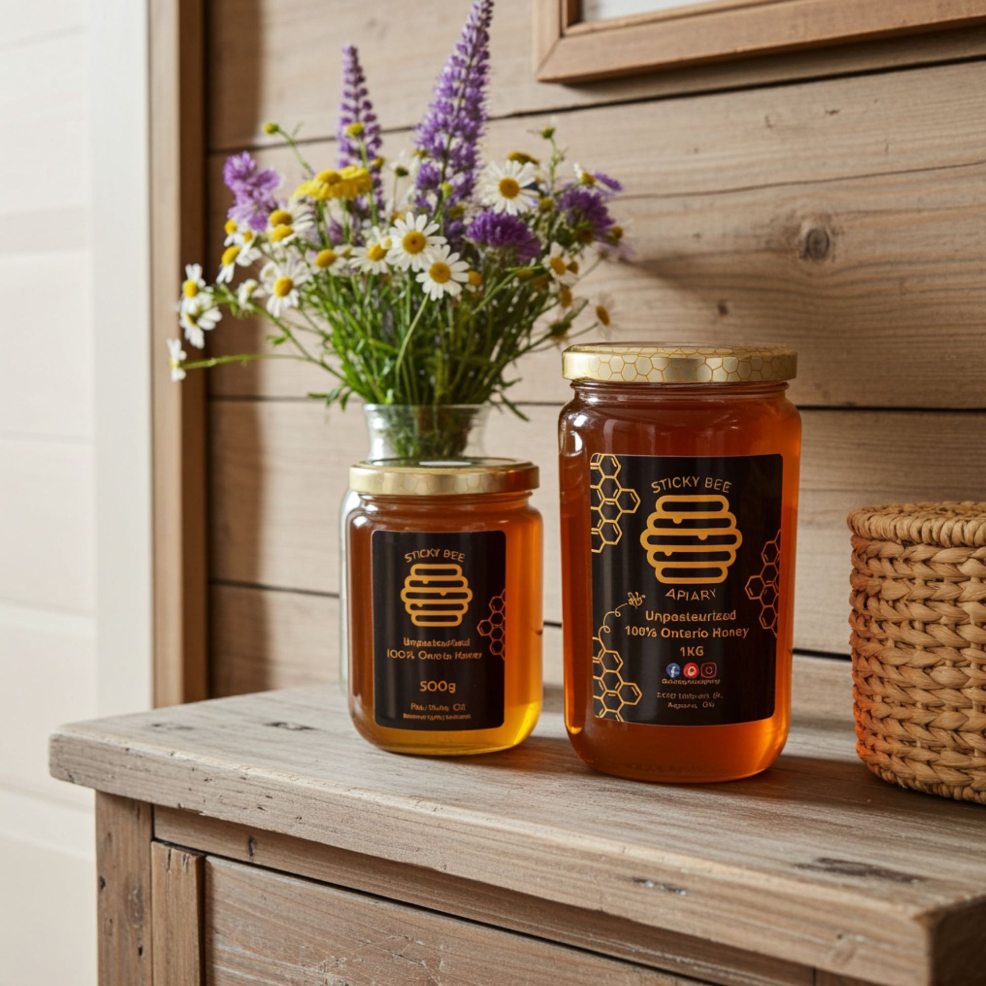 Two jars of honey from Sticky Bee on a wooden surface with a vase of flowers in the background.