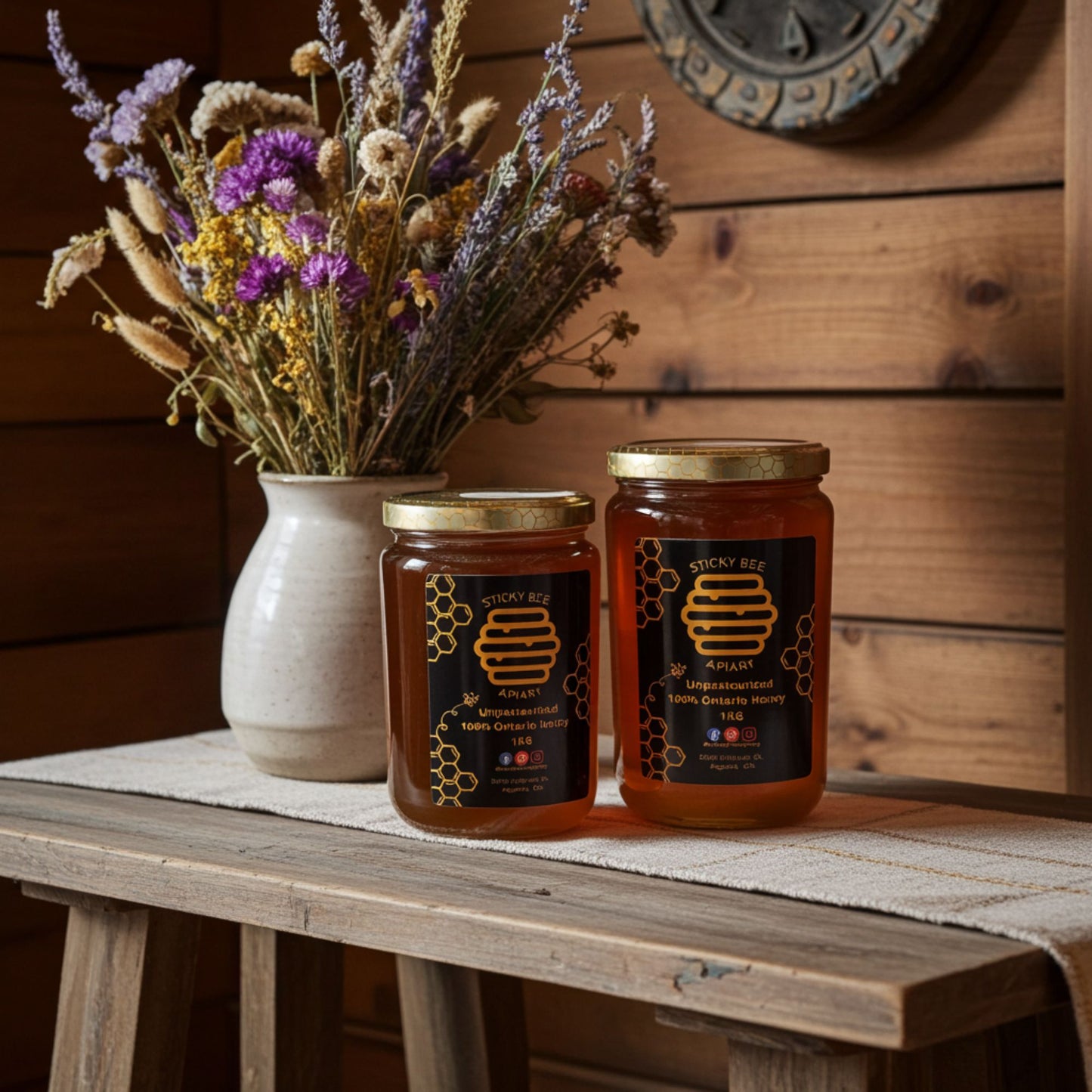 Two jars of honey on a wooden table with a set of flowers to the left.