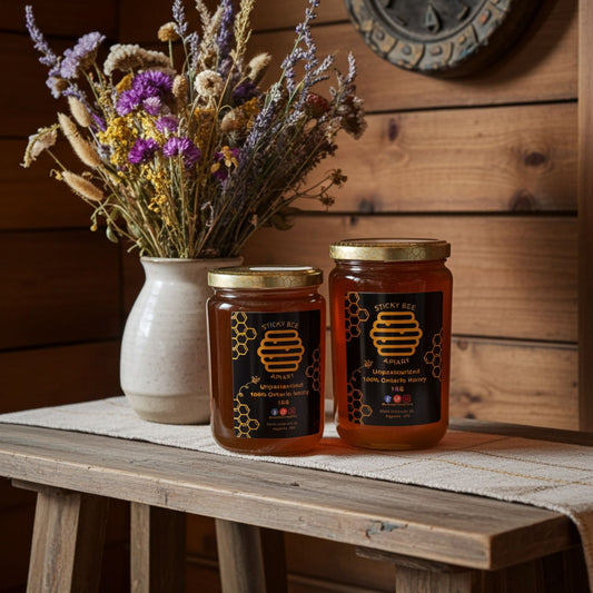 Two jars of honey on a wooden table with a set of flowers to the left.