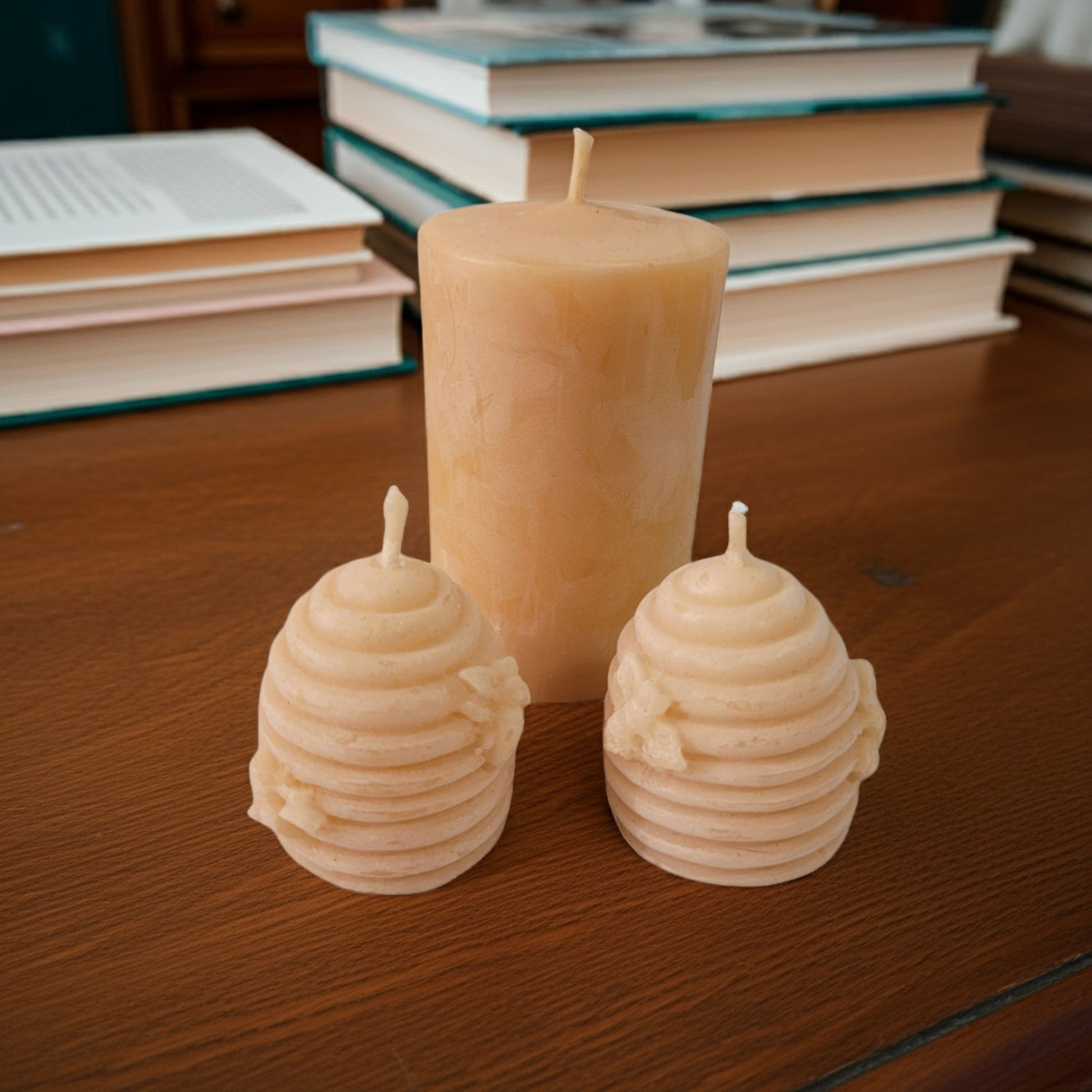 Two beehive shaped beeswax candles and a 2x3 smooth pillar candle on a wooden surface with books in the background.