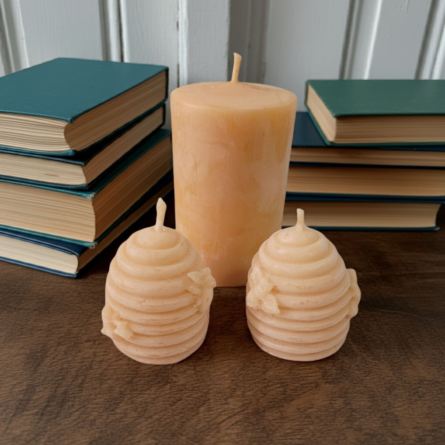 Two beehive-shaped candles and a 2x3 smooth pillar beeswax candle on a wooden surface with books in the background.