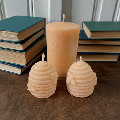 Two beehive-shaped candles and a 2x3 smooth pillar beeswax candle on a wooden surface with books in the background.
