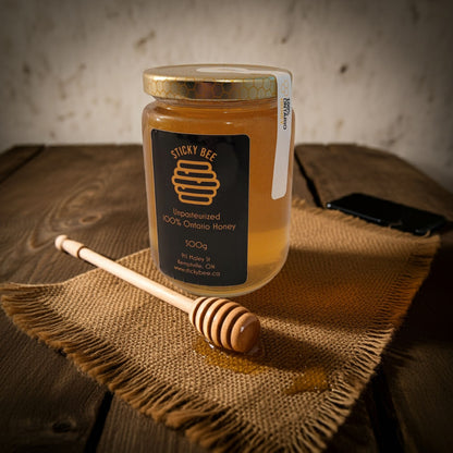Jar of honey with a honey dipper on a textured surface, featuring the 'Sticky Bee' brand.