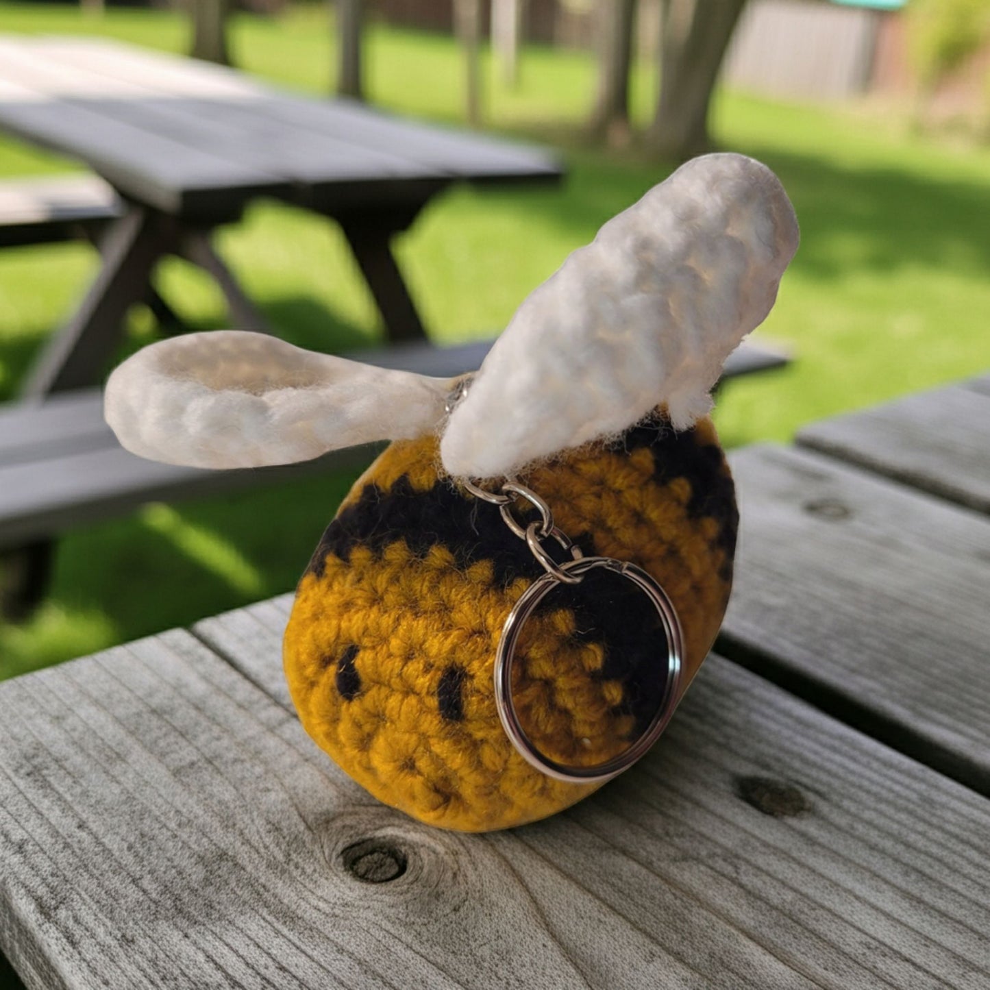 Crocheted bee-shaped keychain on a wooden surface with picnic tables in the background