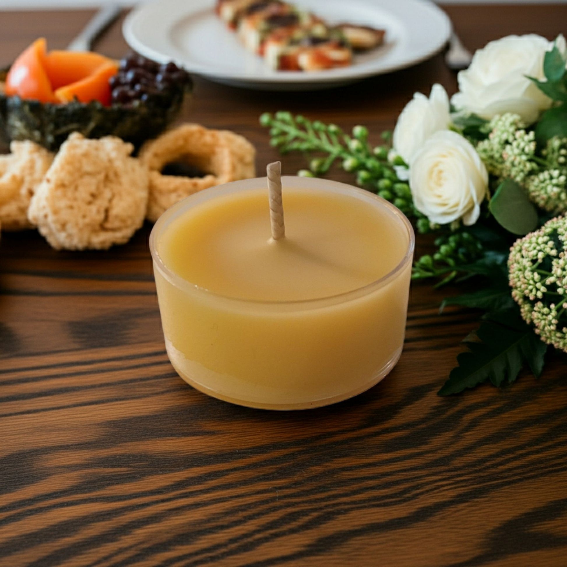Small yellow candle on a wooden table with food and flowers in the background