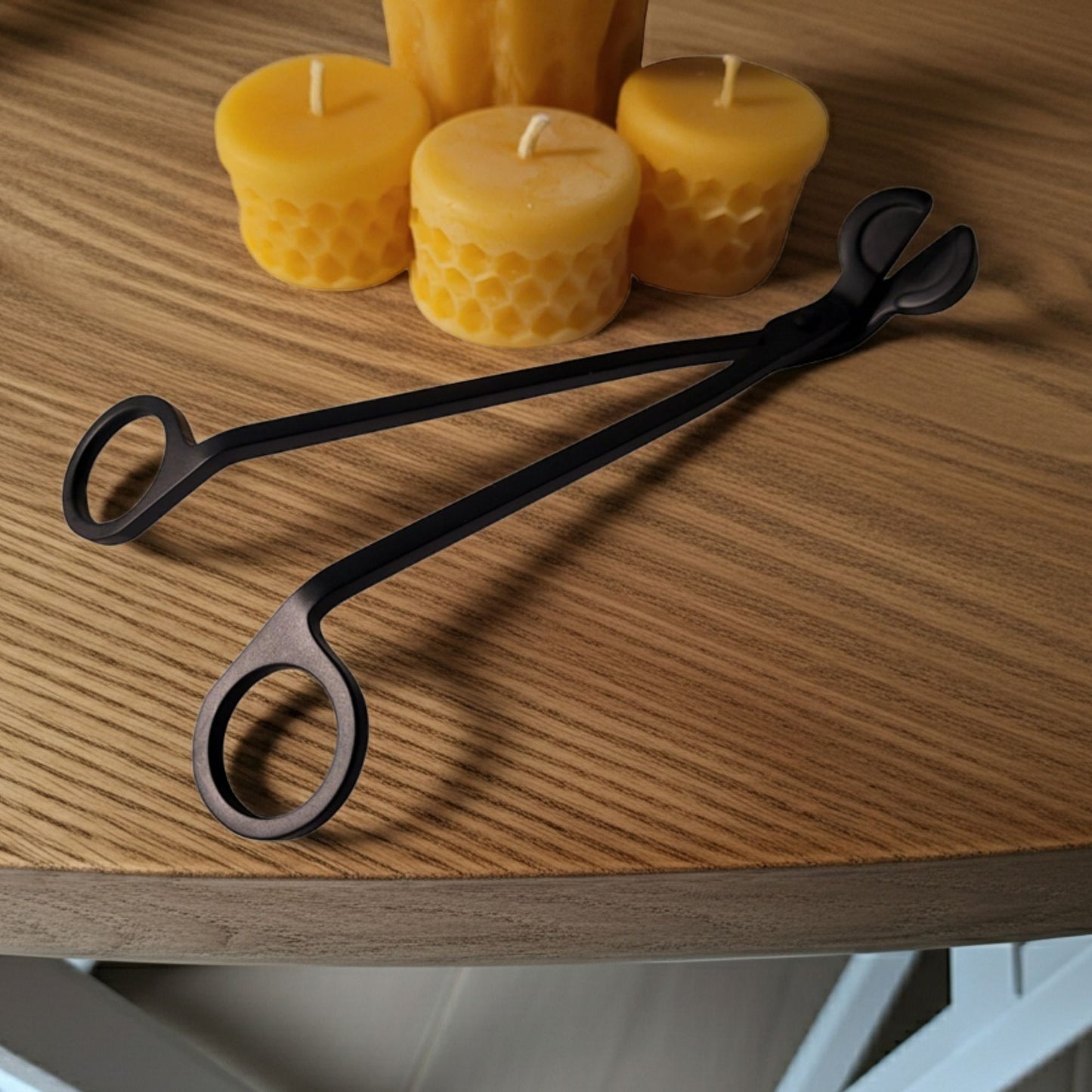 Black wick trimmer on a wooden surface with beeswax candles in the background.
