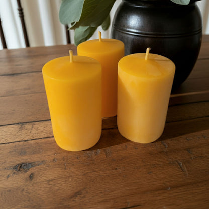 Three yellow candles on a wooden surface with a dark vase and green leaves in the background.