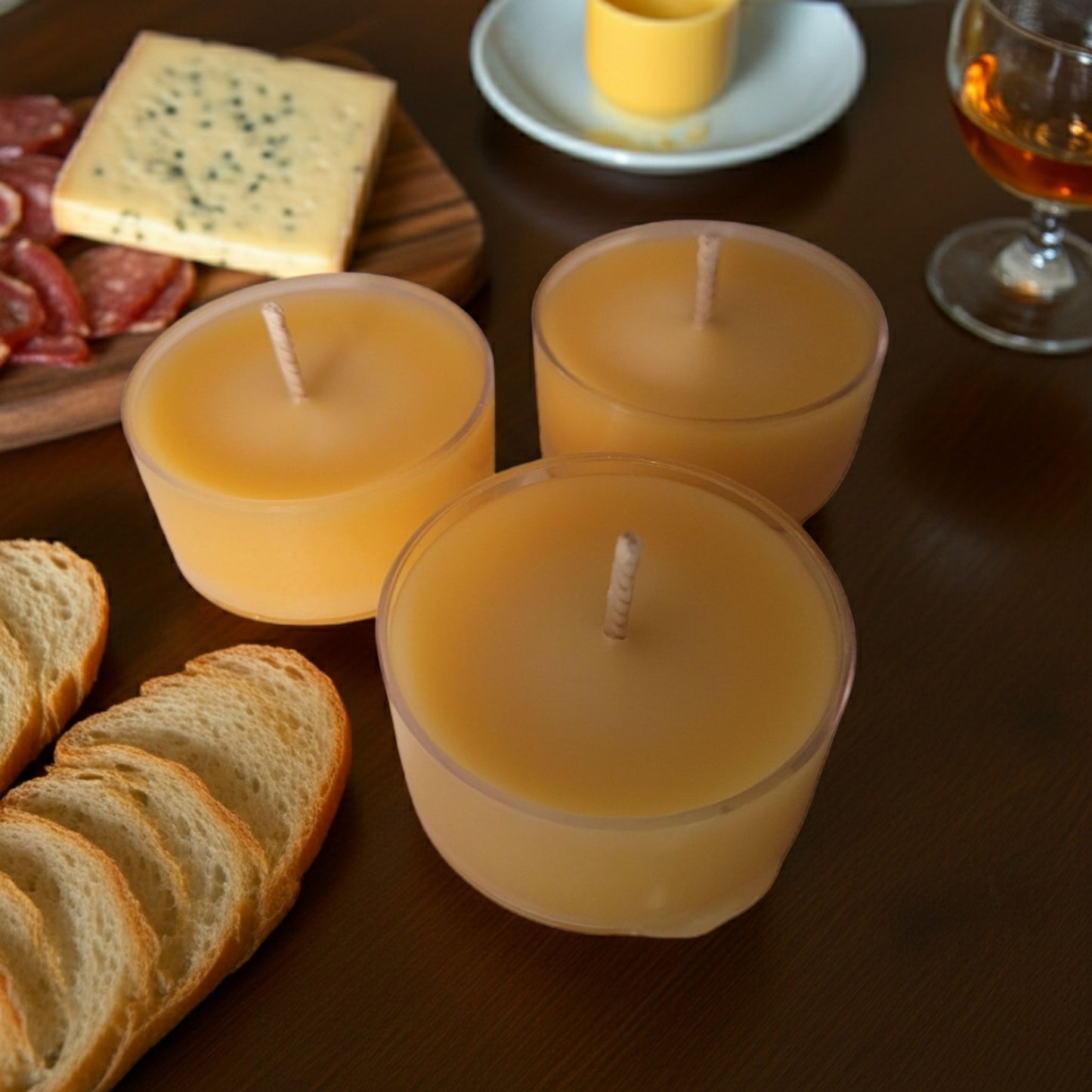 Three small candles on a wooden table with bread, cheese, and a glass of wine in the background.