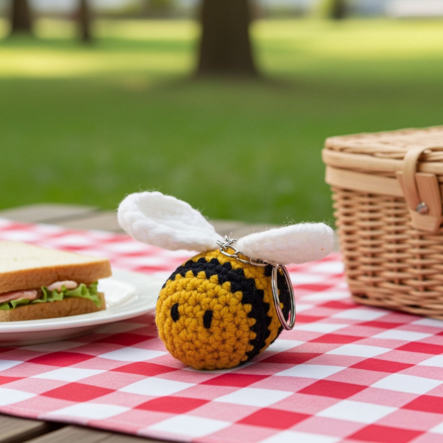 Crocheted bee keychain on a picnic table with a basket and sandwich in the background.