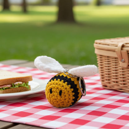 Crocheted bee keychain on a picnic table with a basket and sandwich in the background.